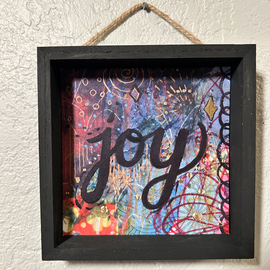 Decorative wall art with the word 'joy' on a colorful background, framed in black and hanging on a textured wall.